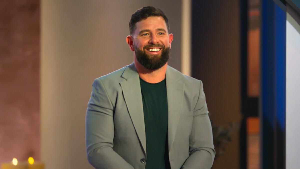 Bearded man in a light gray blazer and dark shirt standing indoors, smiling, with soft lighting and a blurred background featuring neutral walls and dark trim.
