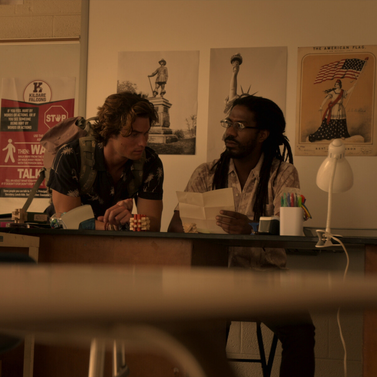Two men sit at a classroom desk, one reading a paper while the other leans in to listen. The room has posters, school supplies, a lamp, and a soft, warm light creating a focused and studious atmosphere.