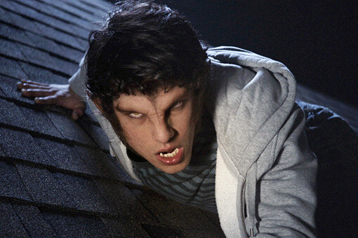 A person with werewolf features crouches on a dark roof at night, wearing a striped shirt and hoodie, with glowing eyes, sharp teeth, and aggressive expression in a dramatic, suspenseful setting.