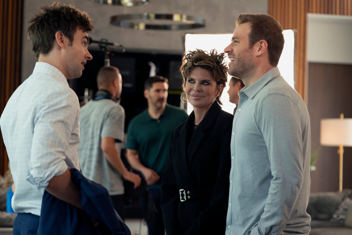 Three people have a conversation in a modern, well-lit living room with crew members, cameras, and equipment visible in the background, suggesting a film or photoshoot set environment.