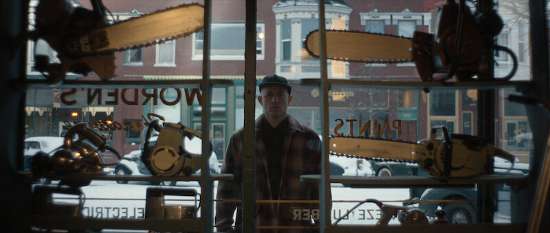 A man in a cap and plaid jacket stands outside a hardware store window, looking in past shelves filled with chainsaws and tools on a snowy day.
