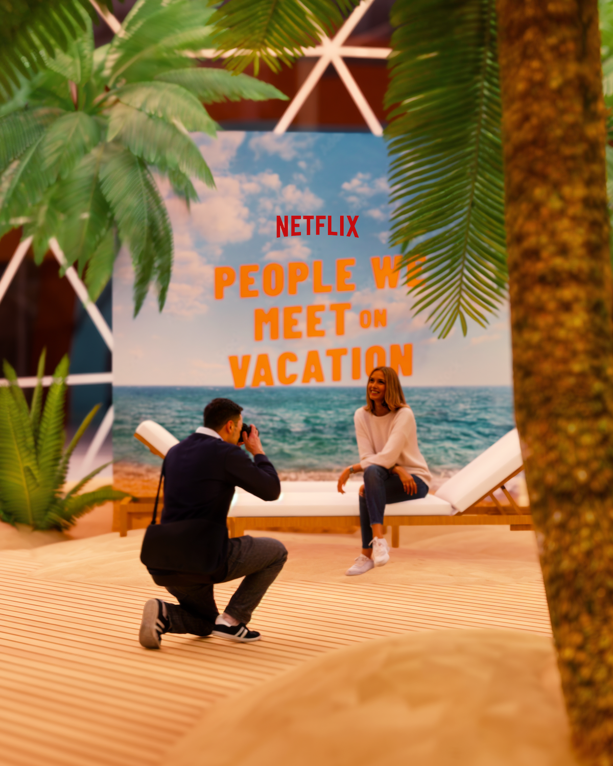 Man taking a photo of a woman sitting on a lounge chair in a tropical-themed indoor set with palm trees, sand, and a Netflix “People We Meet on Vacation” backdrop.