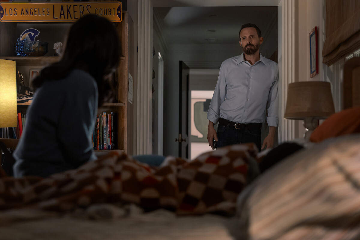 A woman sits on the bed and looks over to a man in a dress shirt who is leaning in the doorway.