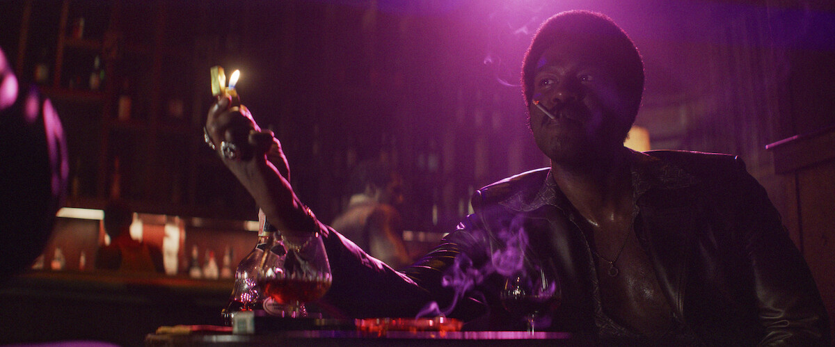 Man in a dimly lit bar, sitting at a table with a drink, lighting a cigarette with a match. Smoke rises in the moody atmosphere, purple and yellow lights create a dramatic, cinematic setting.