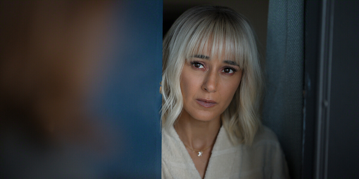 Woman with blond hair and bangs, wearing a light robe, stands in a doorway indoors, looking out with a serious expression, soft lighting and blurred blue background.