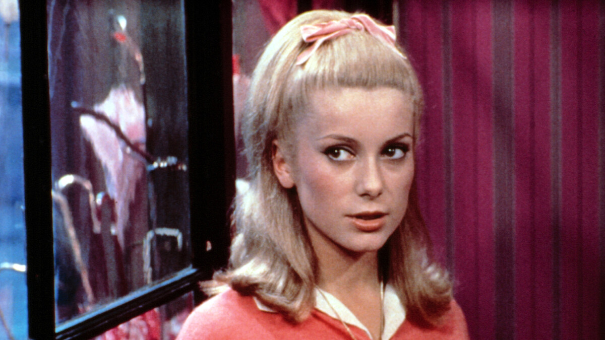 Young woman with blonde hair and pink headband, wearing a pink cardigan, stands indoors near a window. The room has pink striped wallpaper and the lighting is soft, giving a vintage atmosphere.