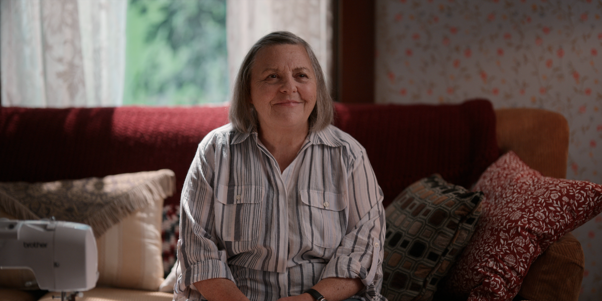Nicola Cavendish as Connie smiles while sitting on a comfortable looking red couch.