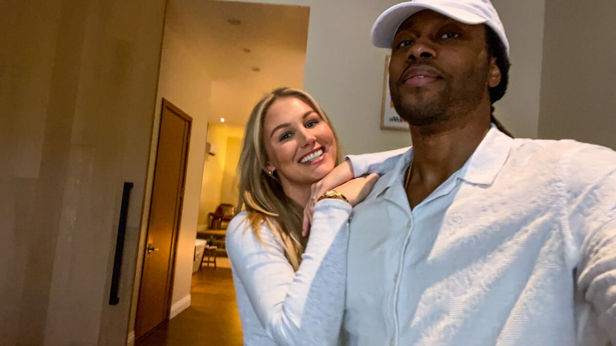 Dr. Vic St. John and Christine Hamilton taking a selfie together inside a warmly lit home, with a hallway and open door in the background. Both are dressed in light-colored clothing; the man is wearing a white cap.