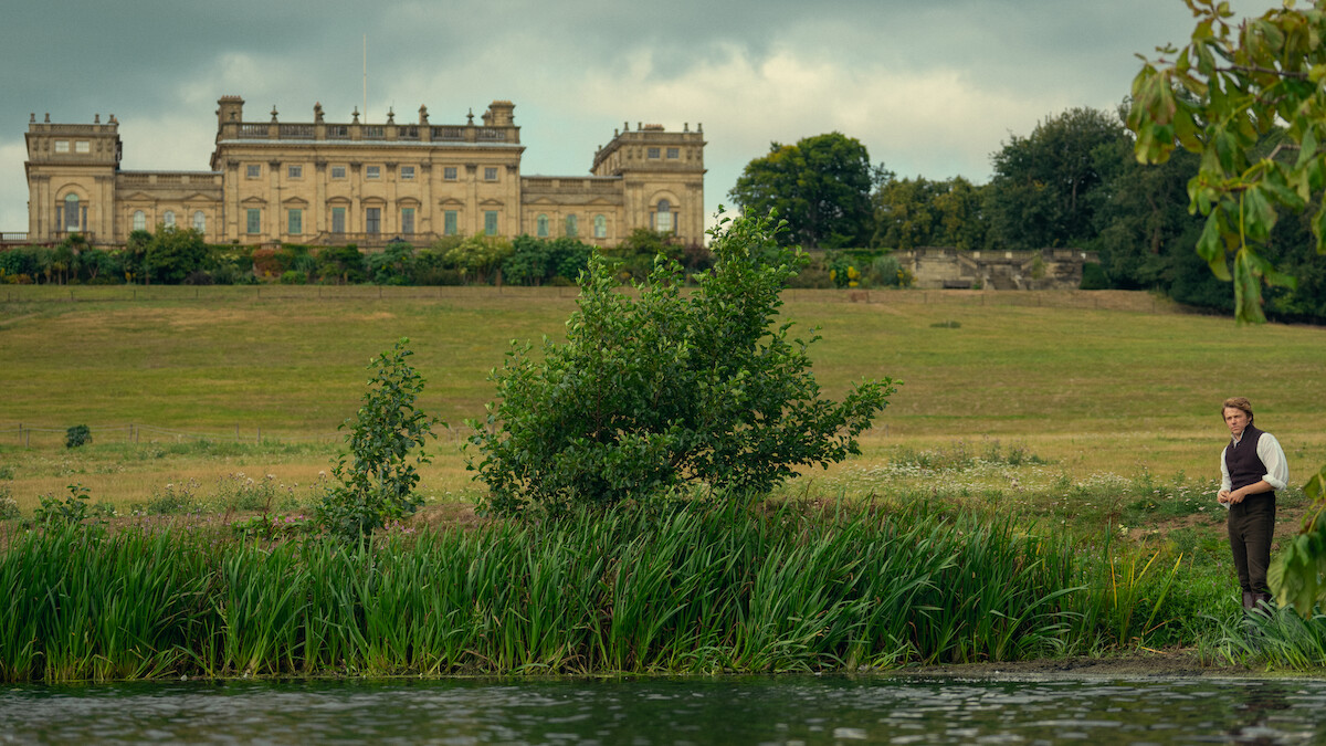 A person stands by a riverbank surrounded by green grass and bushes, with a large historic mansion set on a hill in the background under a cloudy sky.