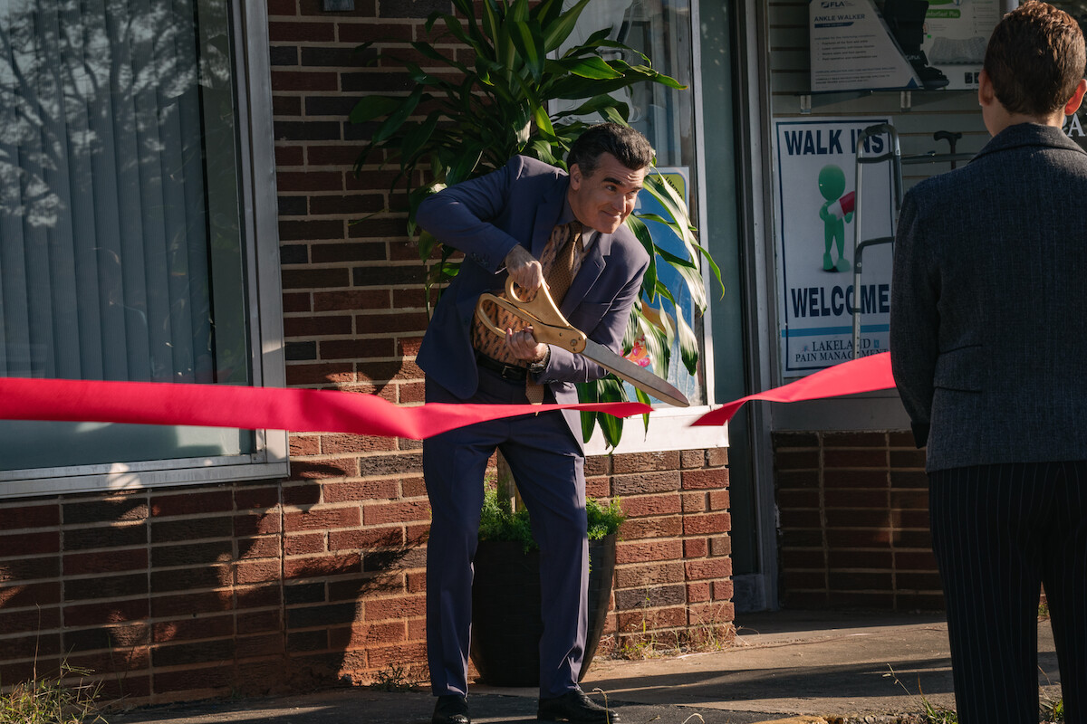 Brian d’Arcy James cuts red tape with a comically large pair of gold scissors in ‘Pain Hustlers.’