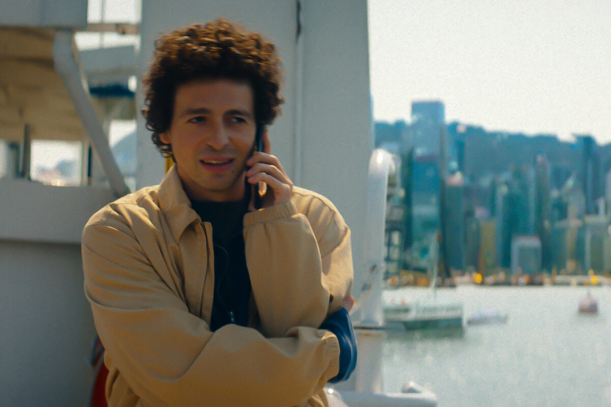 Man with curly hair talking on a cell phone stands outside on a boat or ferry with a city skyline and waterfront in the background on a bright day.