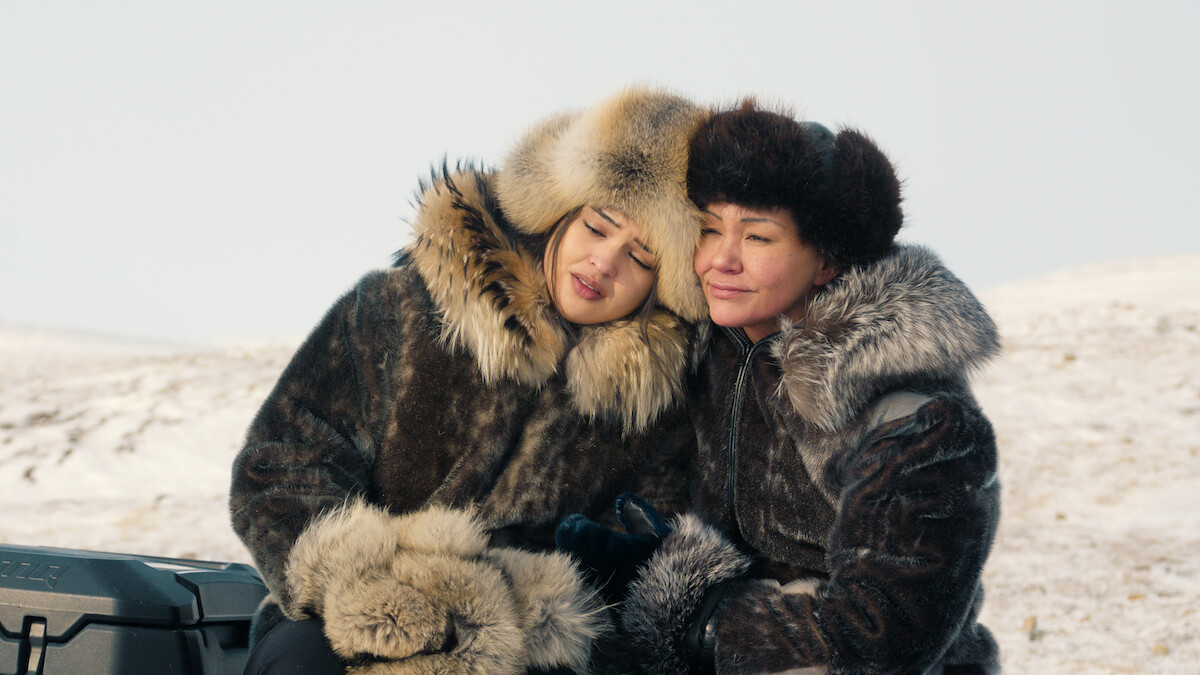 Two people wearing thick fur coats and hats sit close together outdoors, surrounded by a snowy landscape, sharing a peaceful moment in the cold environment.