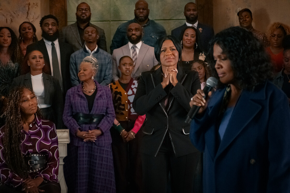 A woman speaks or sings into a microphone in the foreground as a formally dressed group stands behind her, some with solemn or emotional expressions, in an indoor setting resembling a church or gathering space.