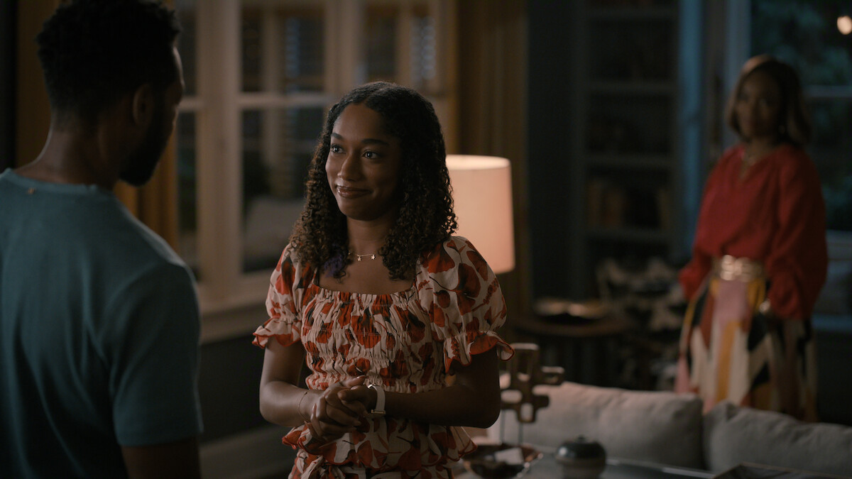 A young woman smiles and talks to a man in a warmly lit living room, with another woman standing in the background near a window and bookshelf. The setting appears cozy and intimate, suggesting a personal or emotional conversation.