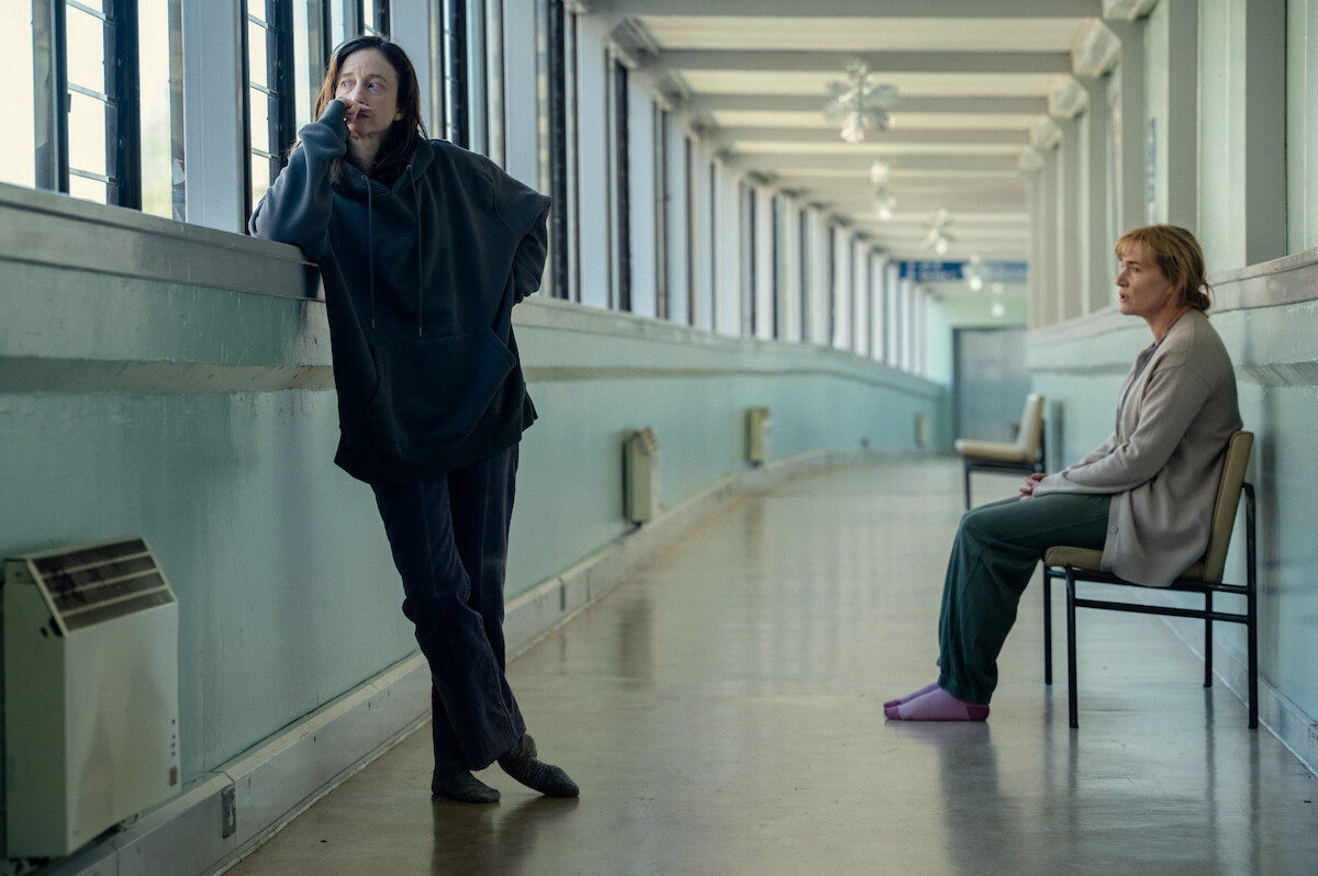 Two women in a long hallway; one stands pensively by the window, the other sits on a chair looking contemplative.