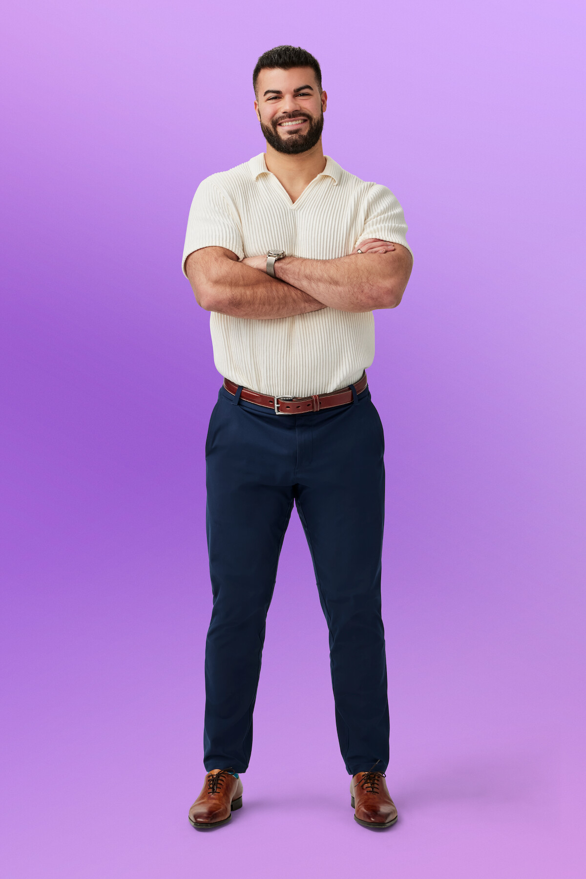 Miguel standing with arms crossed, wearing a short-sleeve shirt, dark pants, and brown shoes, set against a solid light purple background in a studio environment.