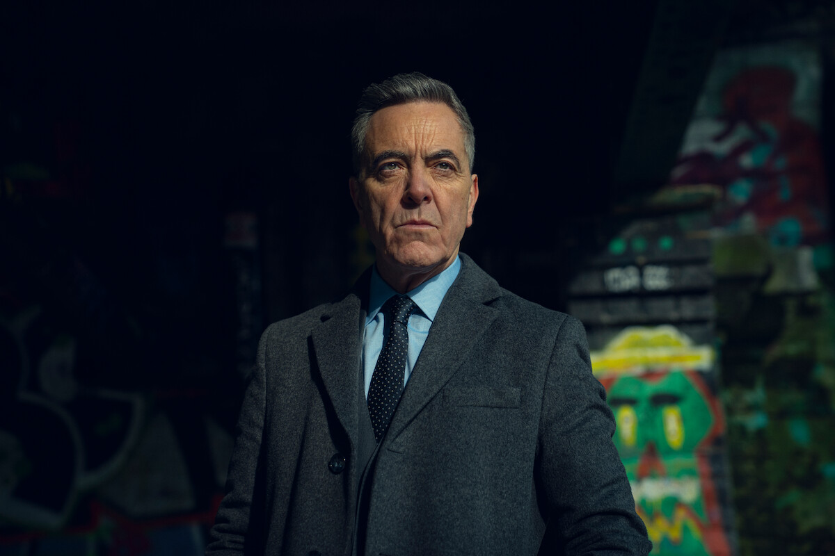 Serious older man in a suit stands confidently in an urban setting with colorful graffiti on walls, creating a gritty, intense atmosphere.
