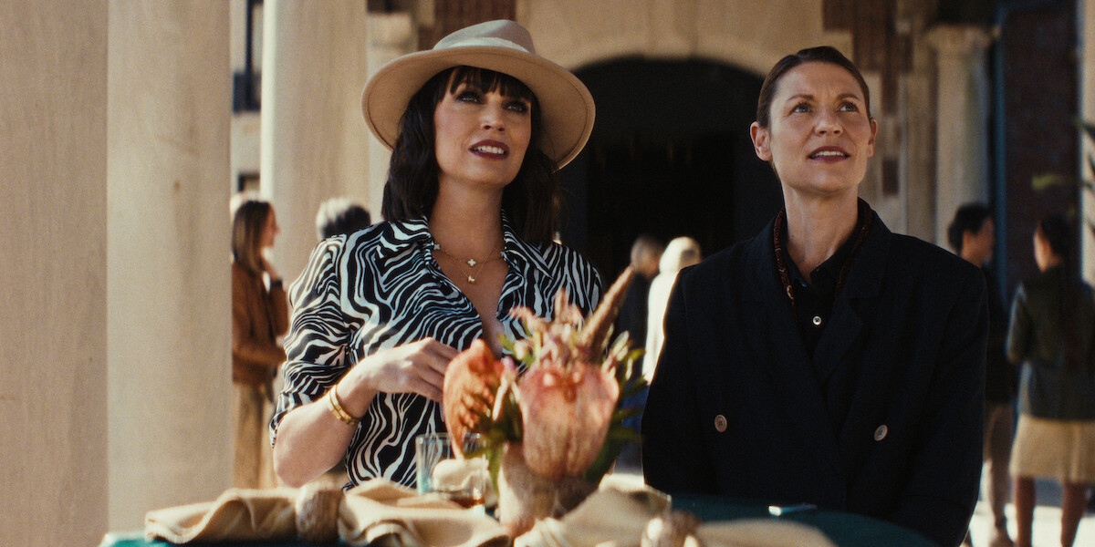 Two women stand at a decorated outdoor table, one in a patterned dress and hat, the other in a dark coat, surrounded by people in a sunlit, elegant setting with a relaxed and cheerful atmosphere.