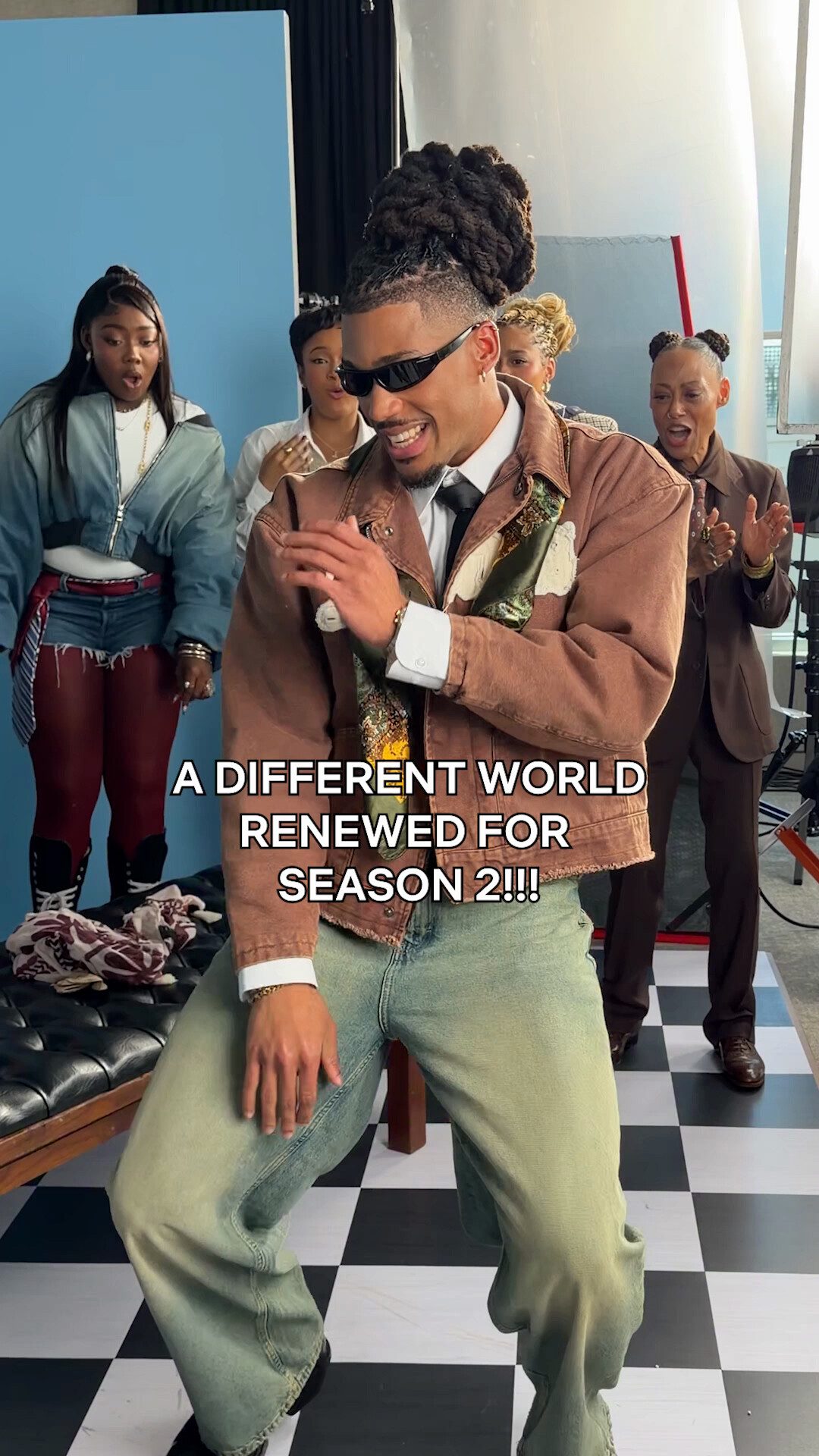 Man dancing joyfully in retro-style outfit, wearing sunglasses, with three people in colorful vintage clothing behind him on a black and white checkered floor, set in a bright studio environment. Text: A Different World renewed for season 2.