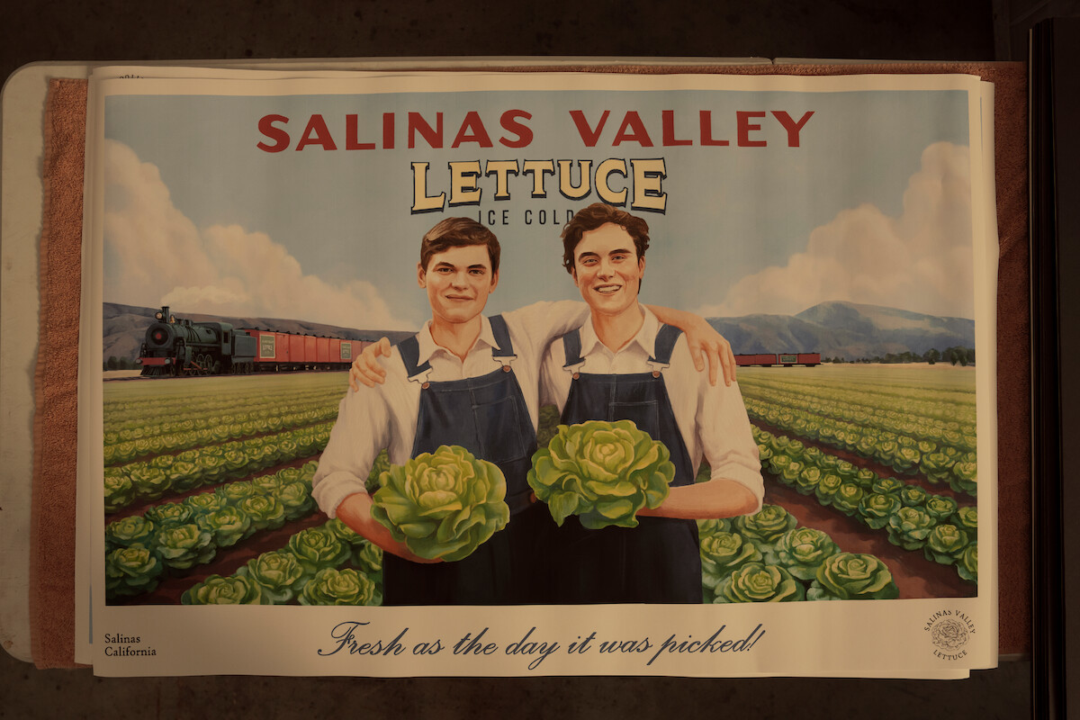 Two smiling men in overalls hold heads of lettuce in a green field with rows of lettuce, a train passing by, and mountains in the background, promoting Salinas Valley lettuce as fresh-picked.