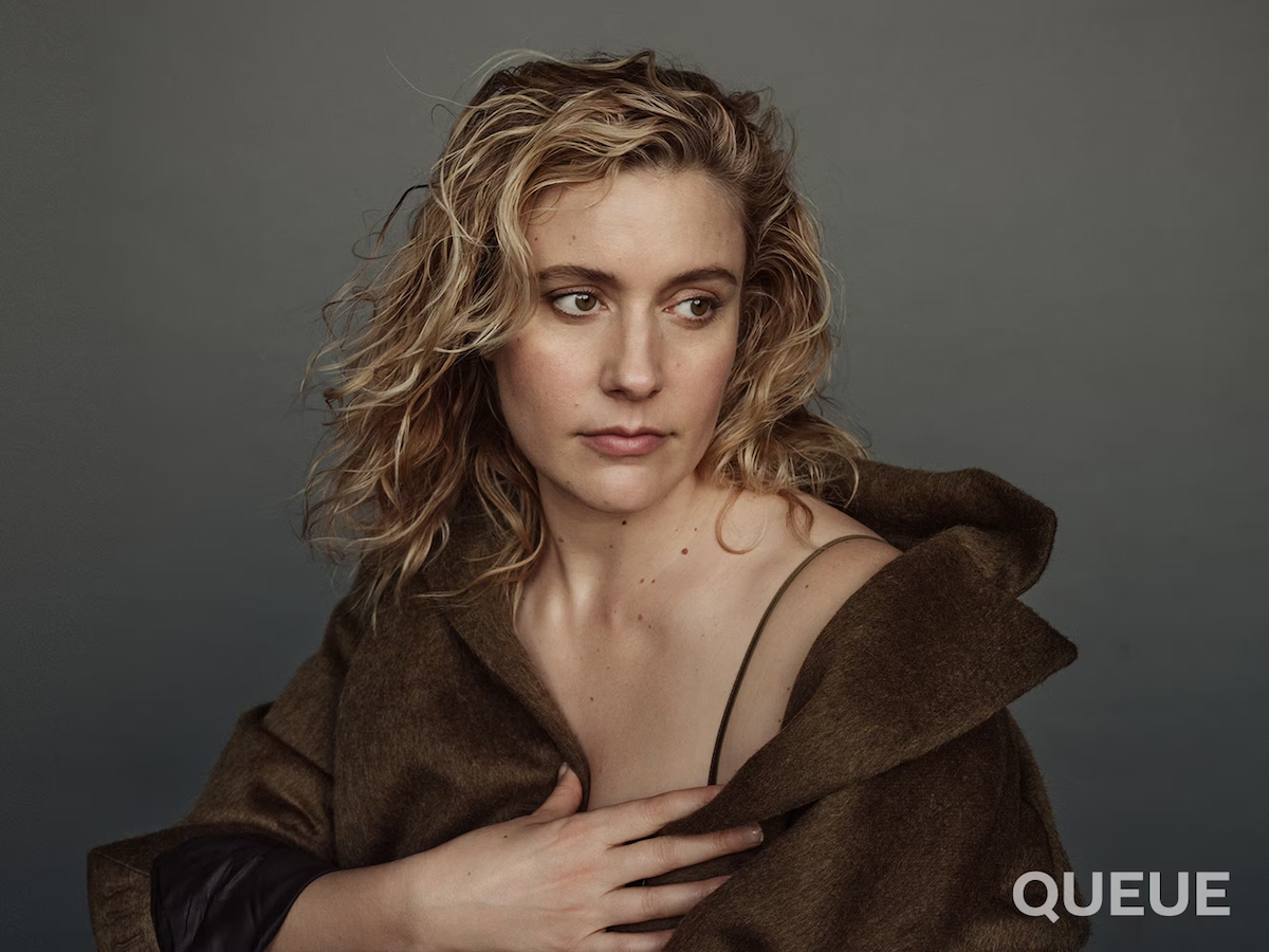Greta Gerwig stares off-camera while wearing a brown coat against a gray backdrop.