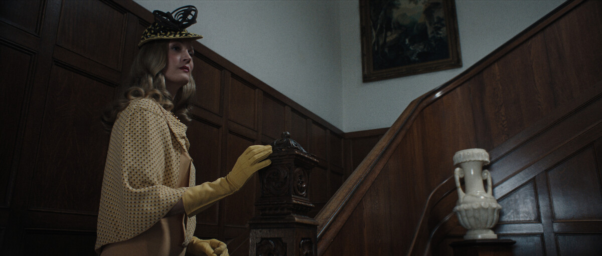 A woman dressed in vintage clothing, including a yellow hat, gloves, and cape, stands at the bottom of a wooden staircase in an elegant, wood-paneled interior.