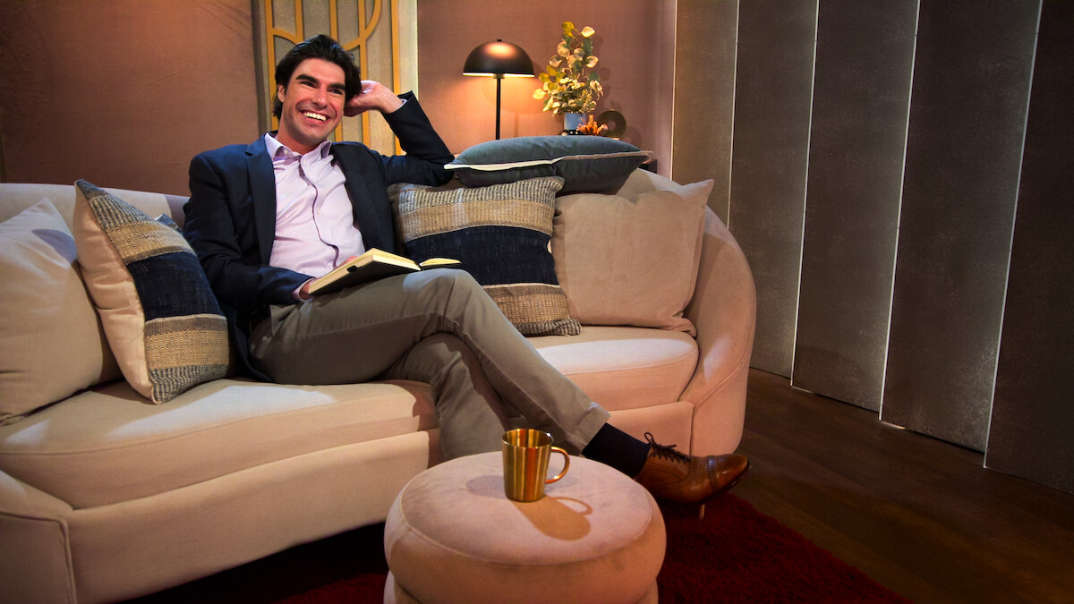 Man in a suit sitting on a modern beige sofa with pillows, holding a book and smiling in a warmly lit room with a lamp, flowers, gold mug on an ottoman, and a red carpet.