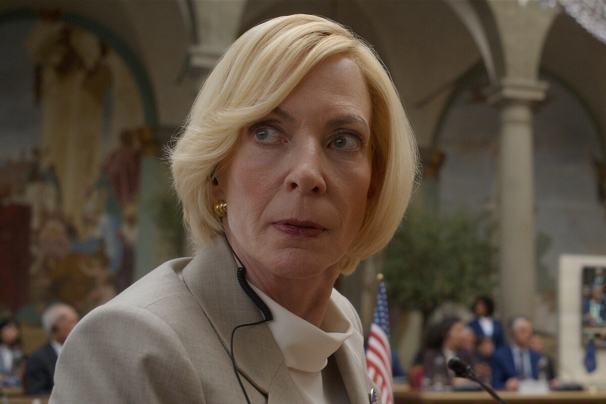 A woman in a light suit with short blonde hair sits in a formal hall with arched columns and murals, looking to the side. An American flag and people in suits are visible in the background, suggesting a government or official setting.