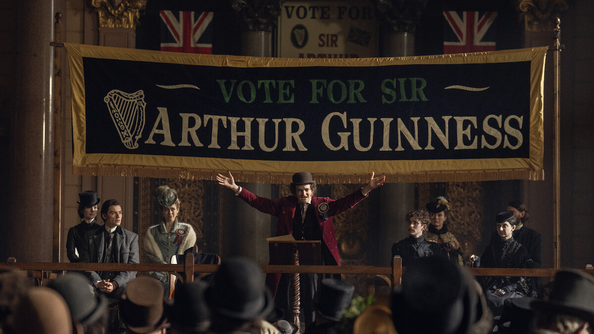 Jack Gleeson as Byron Hedges on a stage under a ‘Vote for Sir Arthur Guinness’ sign in ‘House of Guinness’ Season 1.