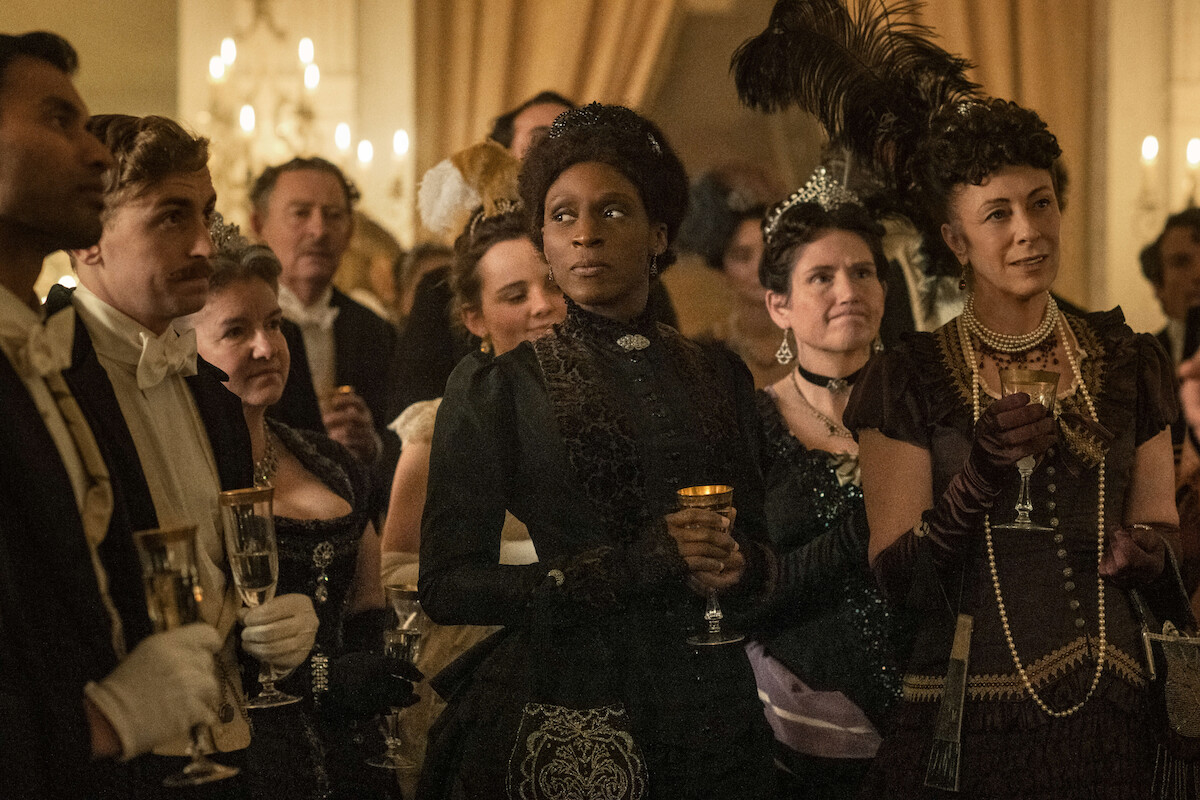A woman in period clothing looks over her shoulder while a group of women and men in regal clothing surround her.