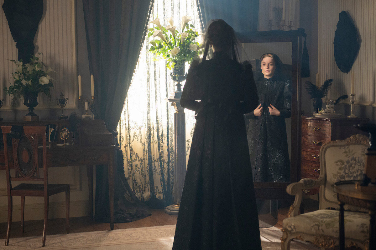 A woman in a dark dress stands in an elegant, vintage room facing a tall mirror, with her reflection visible. The room is softly lit by daylight through lace curtains, surrounded by classic furniture, flowers, and ornate decor.