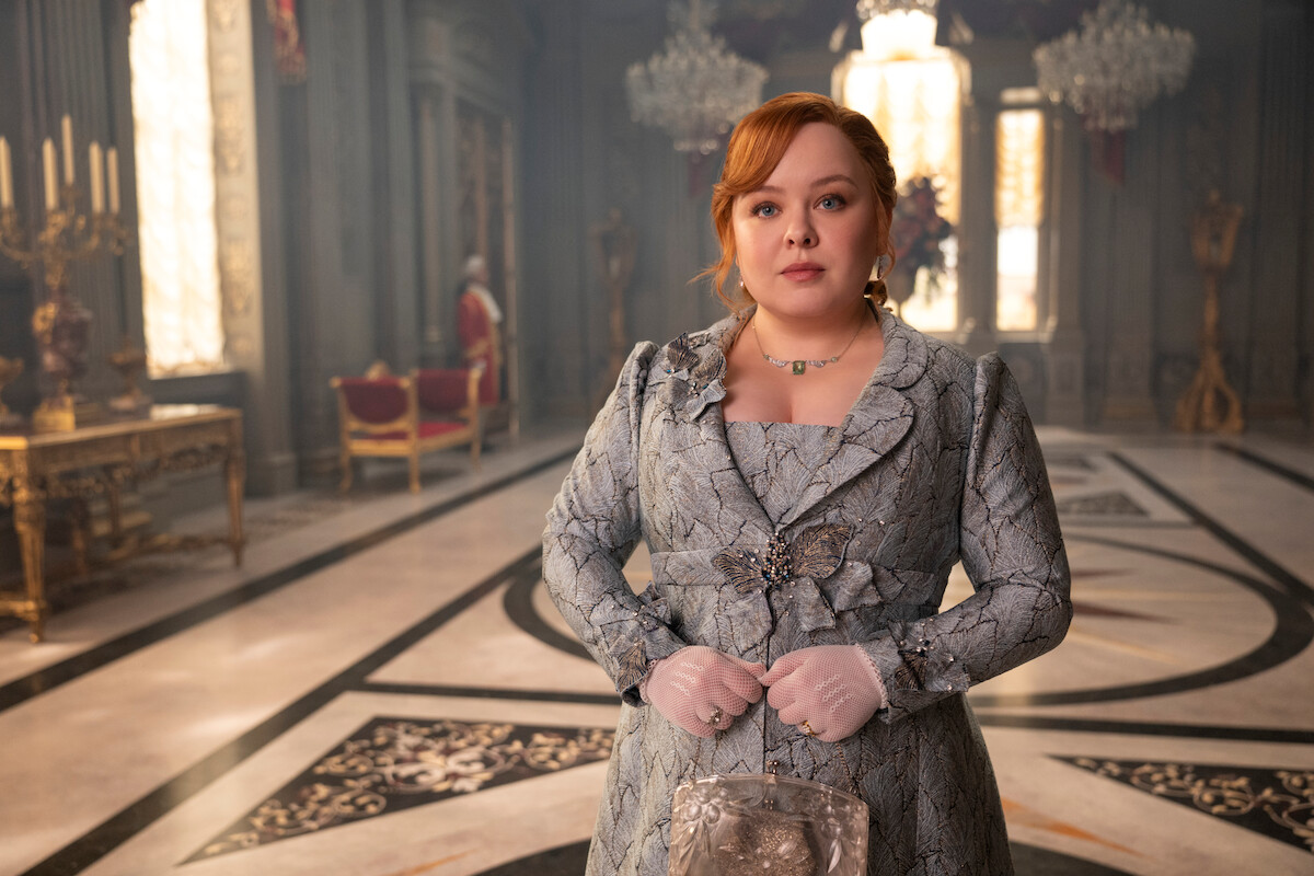 Mulher com um vestido ornamentado de época em pé em um salão elegante de palácio com lustres, piso de mármore e decoração luxuosa. Tem um olhar tranquilo e as mãos fechadas com luvas.