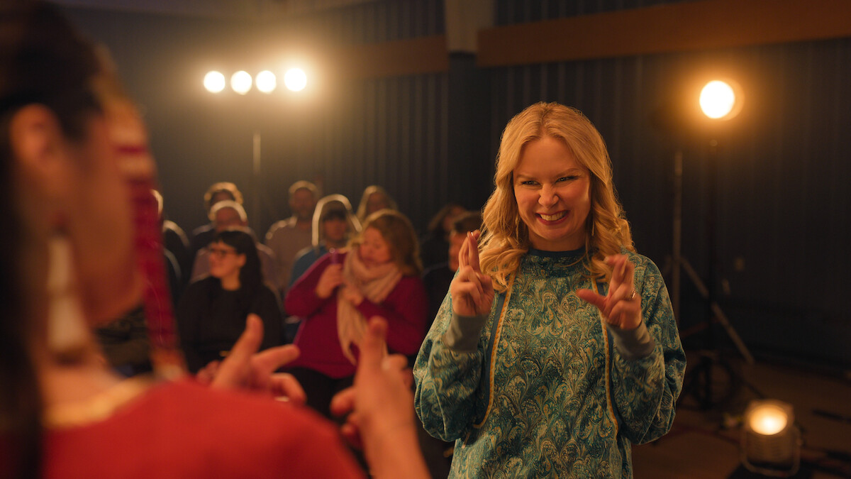 A woman in front making hand gestures, possibly signing, smiles at another person in a warmly lit room with an audience watching, stage lights visible in the background, creating a lively and engaged atmosphere.