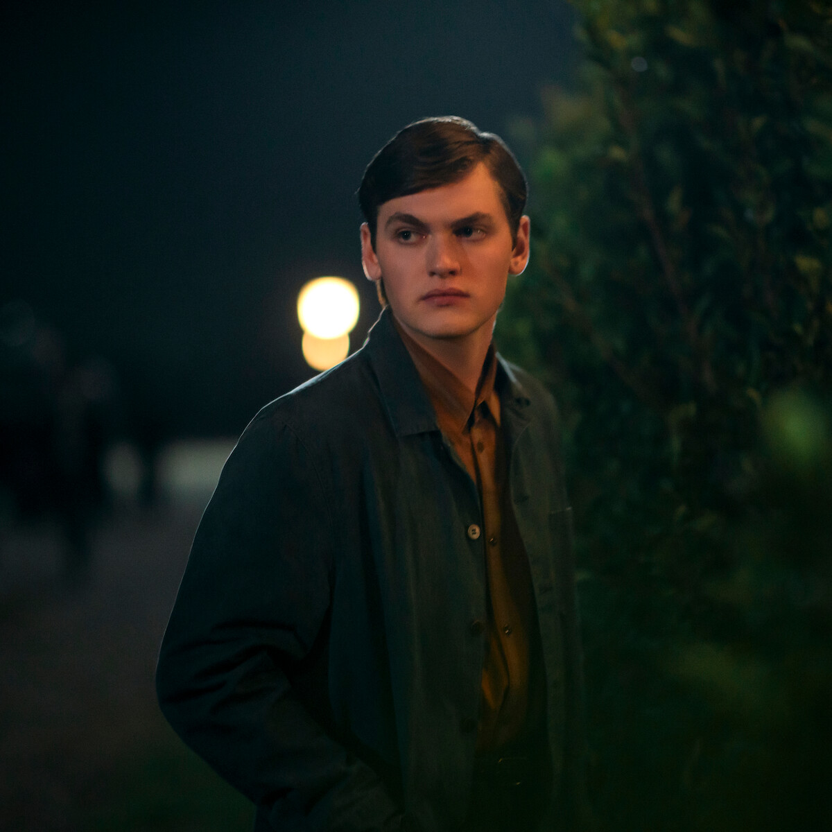 Joe Anders as Aron Trask in a dark jacket stands outdoors at night near a bush, with blurred lights and people in the background, creating a moody and mysterious atmosphere.