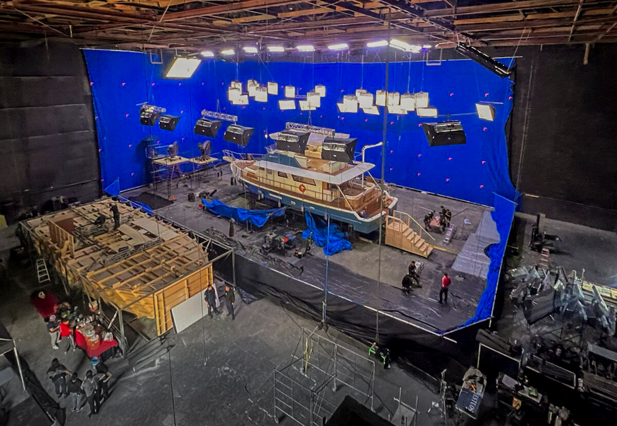 Um grande barco pintado nas cores azul e branco está no meio de um set de filmagem, cercado por pedaços de lona e holofotes de iluminação