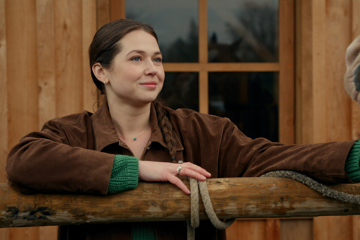 Woman in a brown jacket leans on a wooden fence outside a wooden building, smiling and looking off to the side, with a window and some trees in the background.