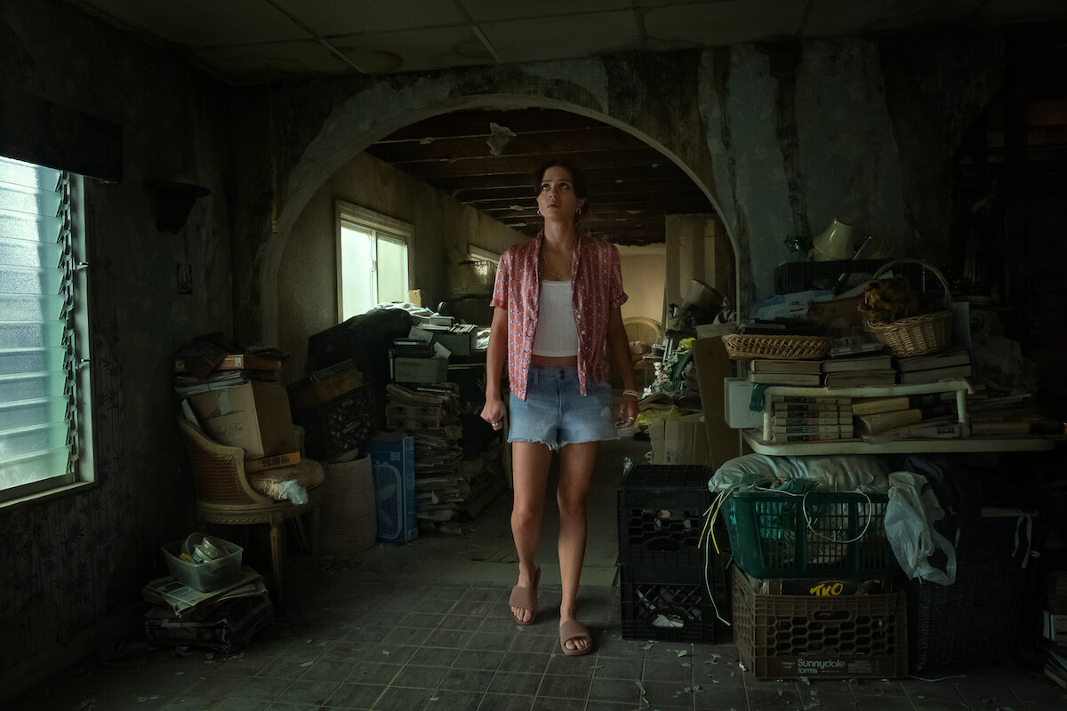 Woman in casual clothes stands barefoot in a cluttered, dimly lit room filled with old furniture, stacked books, and crates, with peeling walls and a broken ceiling, evoking a sense of neglect and disarray.