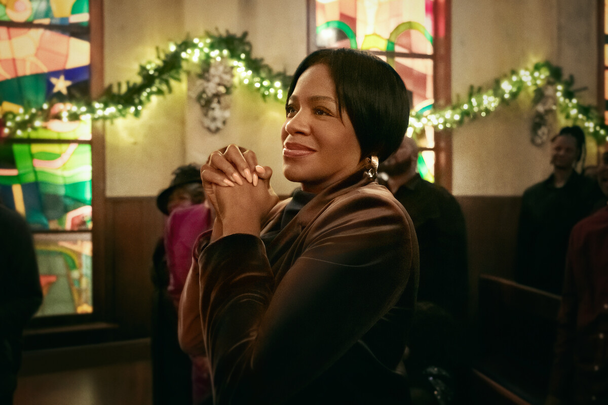 A woman standing in a church, hands clasped in prayer, wearing a brown jacket. Stained glass windows, green holiday garlands, and other people are visible in the background, creating a warm, festive atmosphere.