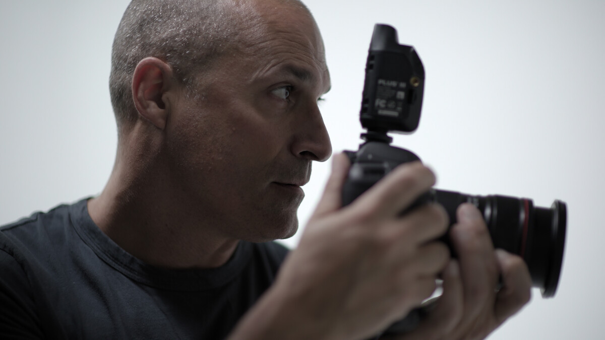 A bald man in a dark shirt holding a camera with a flash attachment, focusing intently in a well-lit, minimal studio environment with a plain light background.