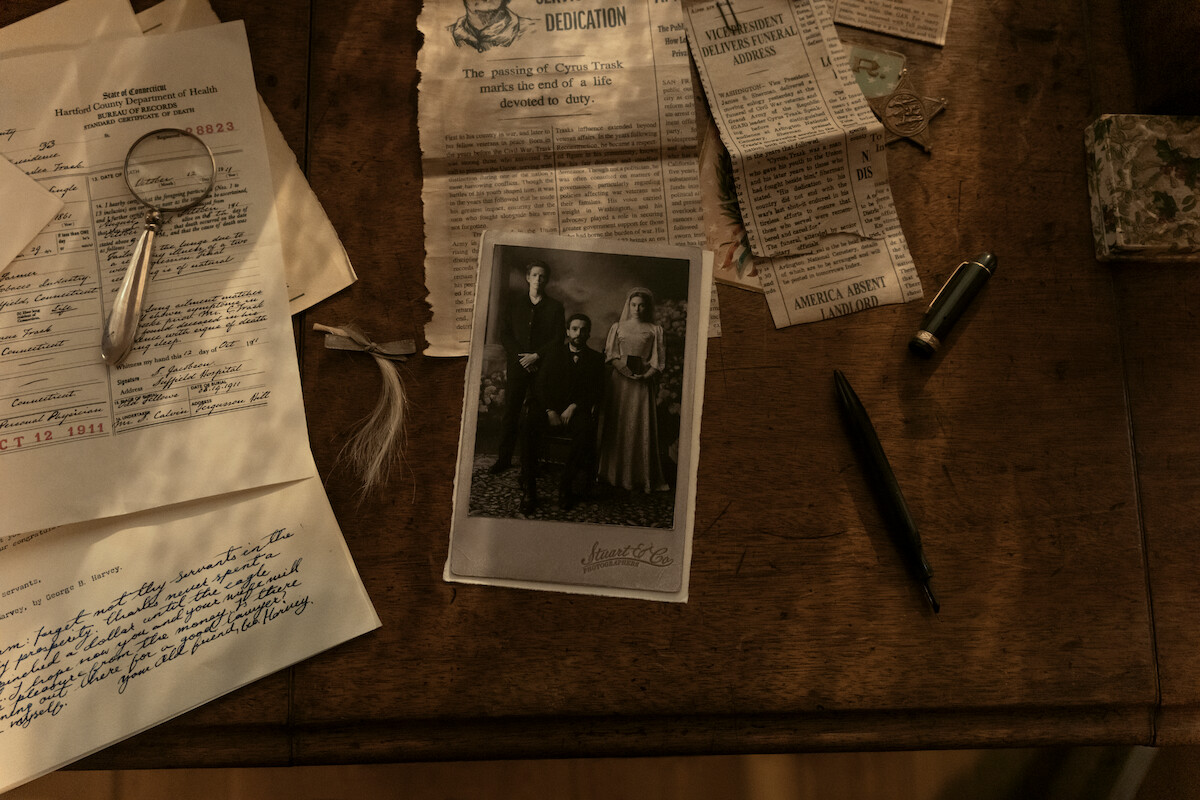 Old sepia photograph, handwritten letters, magnifying glass, pen, and newspaper clippings spread out on a wooden desk, suggesting investigation or family history research in a vintage setting.