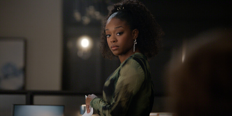 A young woman with curly hair stands indoors in an office-like setting, looking over her shoulder with a serious, contemplative expression. The lighting is soft and the background is blurred, adding to the thoughtful mood.