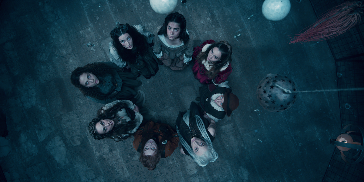 Eight people in medieval-style clothing stand in a circle on a stone floor, looking up toward the camera, surrounded by hanging spherical objects in a dimly lit, mysterious room.