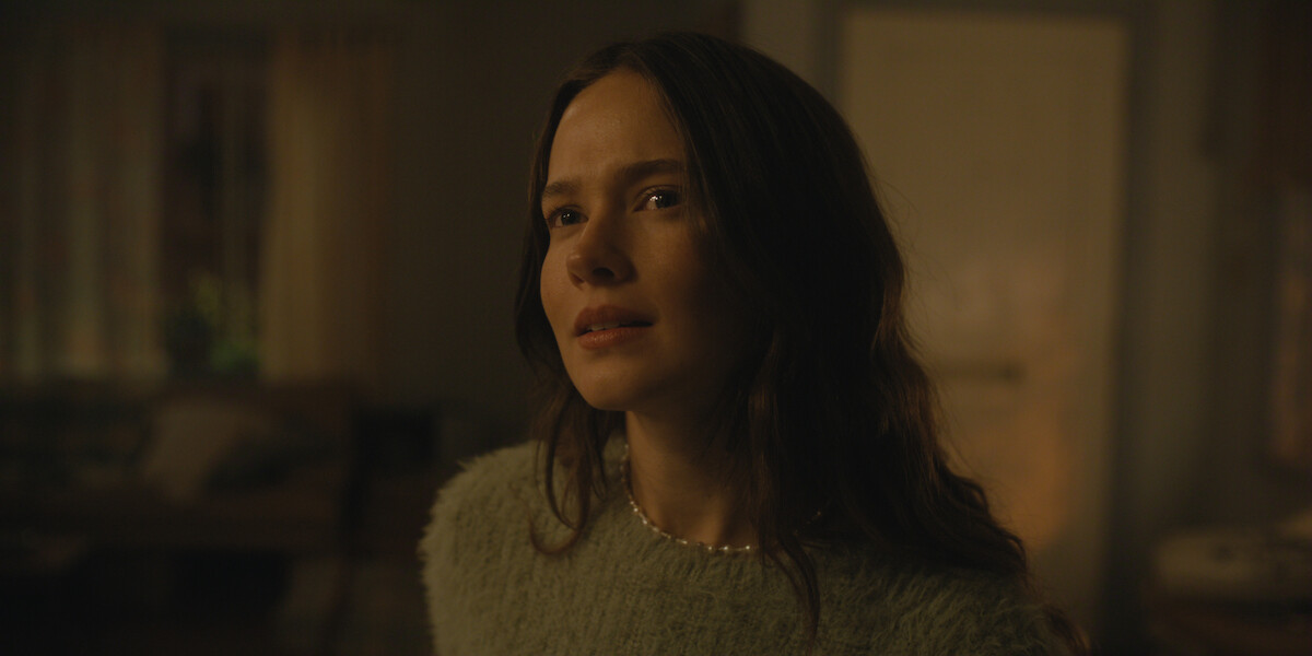 Woman with long brown hair in a green fuzzy sweater stands indoors in a dimly lit cozy room with soft, warm lighting, blurred furniture, and curtains visible in the background.
