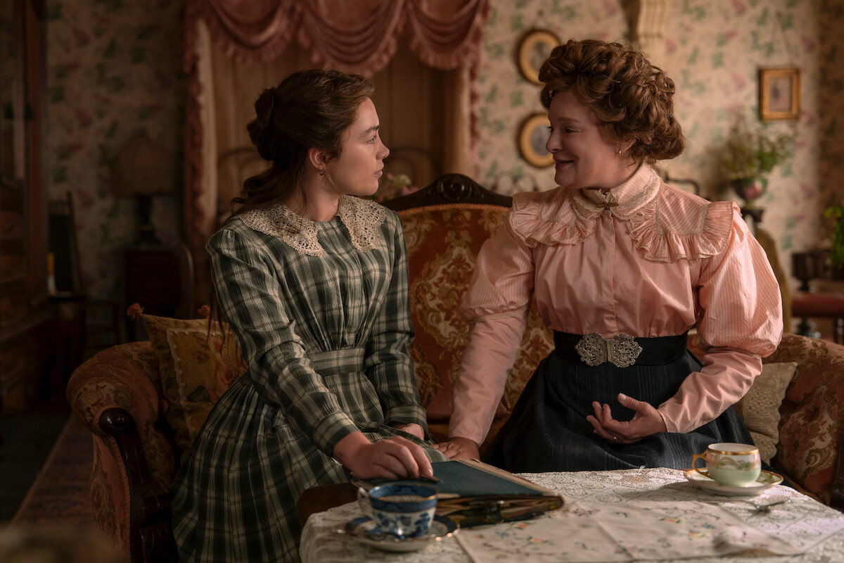 Two women in period clothing sit on a vintage sofa in a richly decorated Victorian-era living room, talking and smiling over tea at a small table with a floral tablecloth and porcelain cups.
