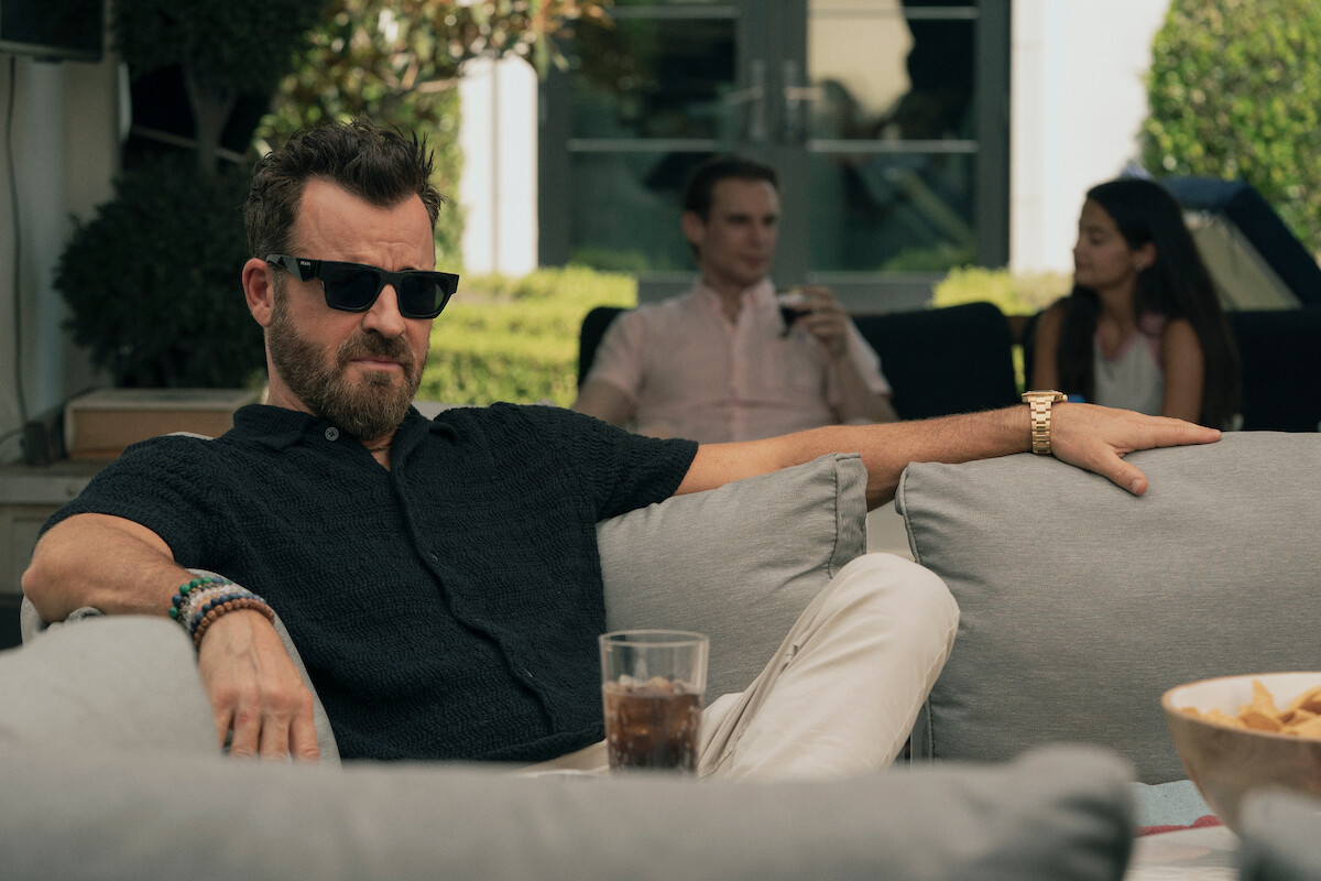 Man in sunglasses relaxing on outdoor couch with a drink, casually dressed, bowl of snacks in foreground; two people talking in the background, modern patio setting with greenery and sunlight.