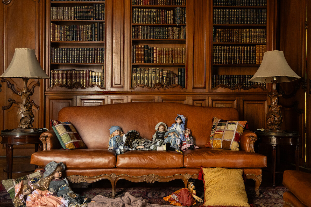 Uma sala ampla com paredes de madeira tem um grande sofá de couro marrom no centro com bonecas sobre sua almofada central. No fundo, uma estante repleta de livros e duas luminárias completam o cenário