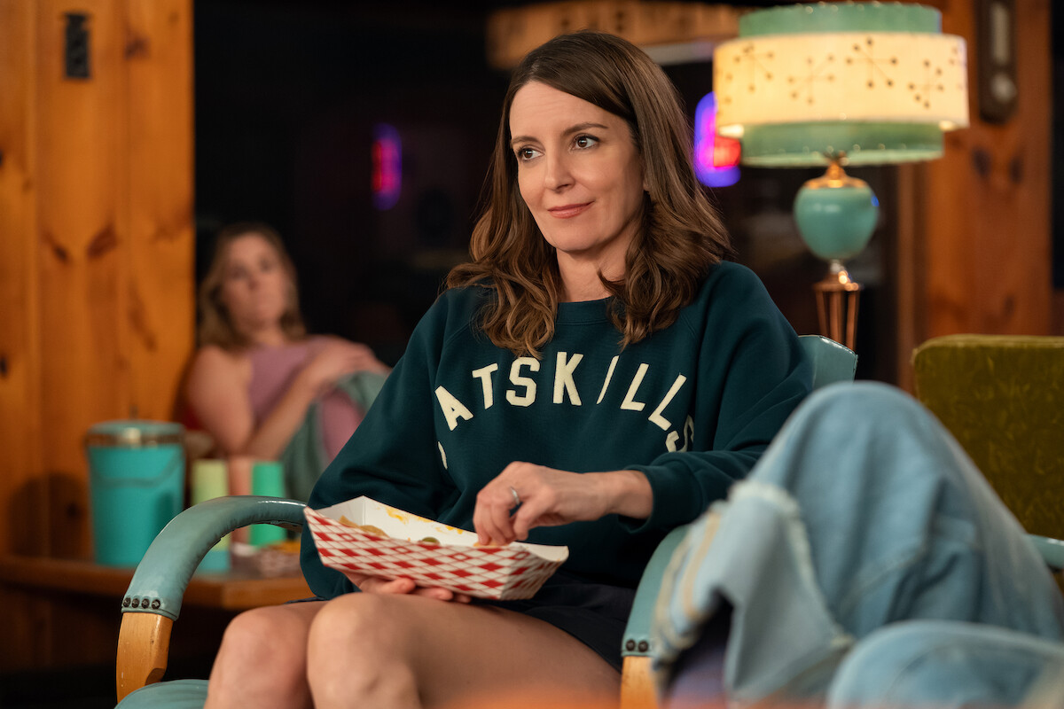 Woman sitting on a retro teal chair indoors, eating from a paper tray and wearing a dark Catskills sweatshirt; cozy, wood-paneled room with a vintage lamp and another person relaxing in the background.