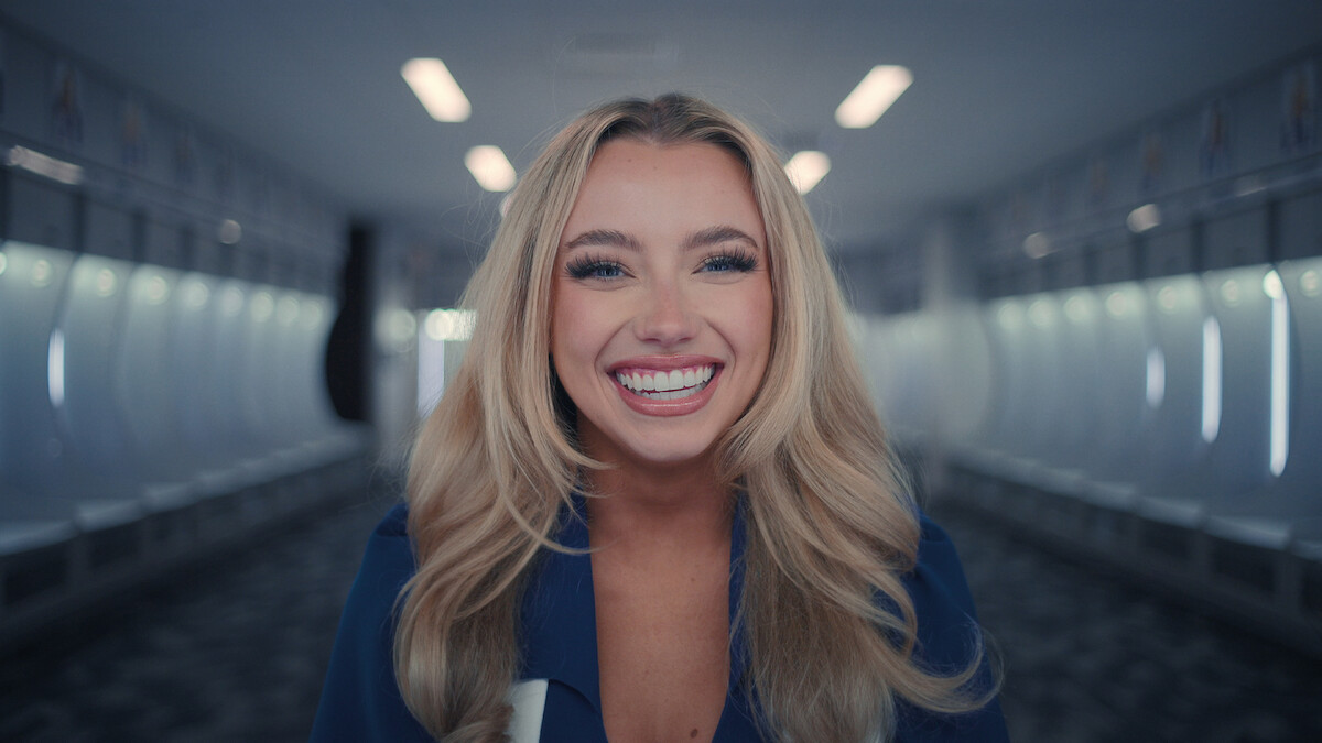 Mulher sorridente olha diretamente para a câmera em um estádio coberto vazio usando o uniforme do DCC, com luzes acesas e arquibancadas vazias ao fundo.