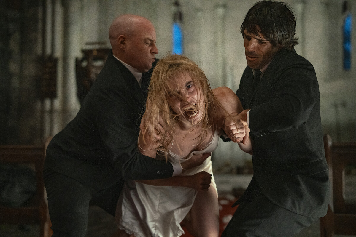 Two men in black suits restrain a distressed woman in a white dress inside a dimly lit church, creating a tense and dramatic atmosphere.