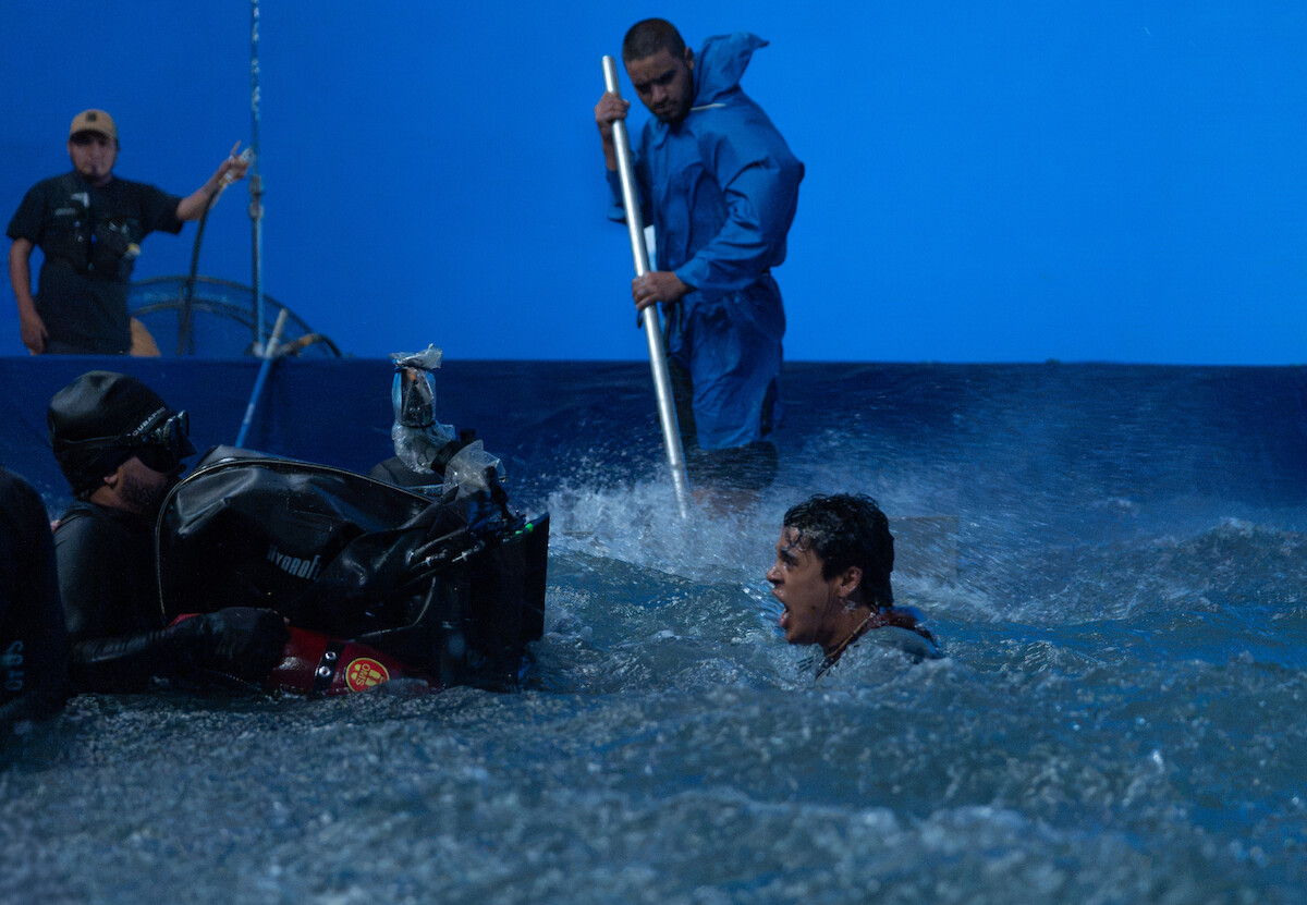 Um homem está dentro da água, com apenas a cabeça para fora. É possível ver que ele está em um tanque de água dentro de um set de filmagem, com uma parede azul no fundo